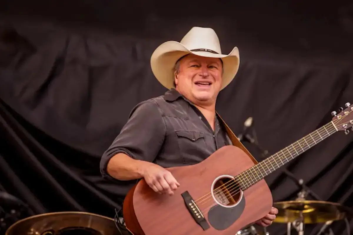 Mark Chesnutt performing on stage, smiling while holding an acoustic guitar, wearing a white cowboy hat and dark shirt under concert lighting.