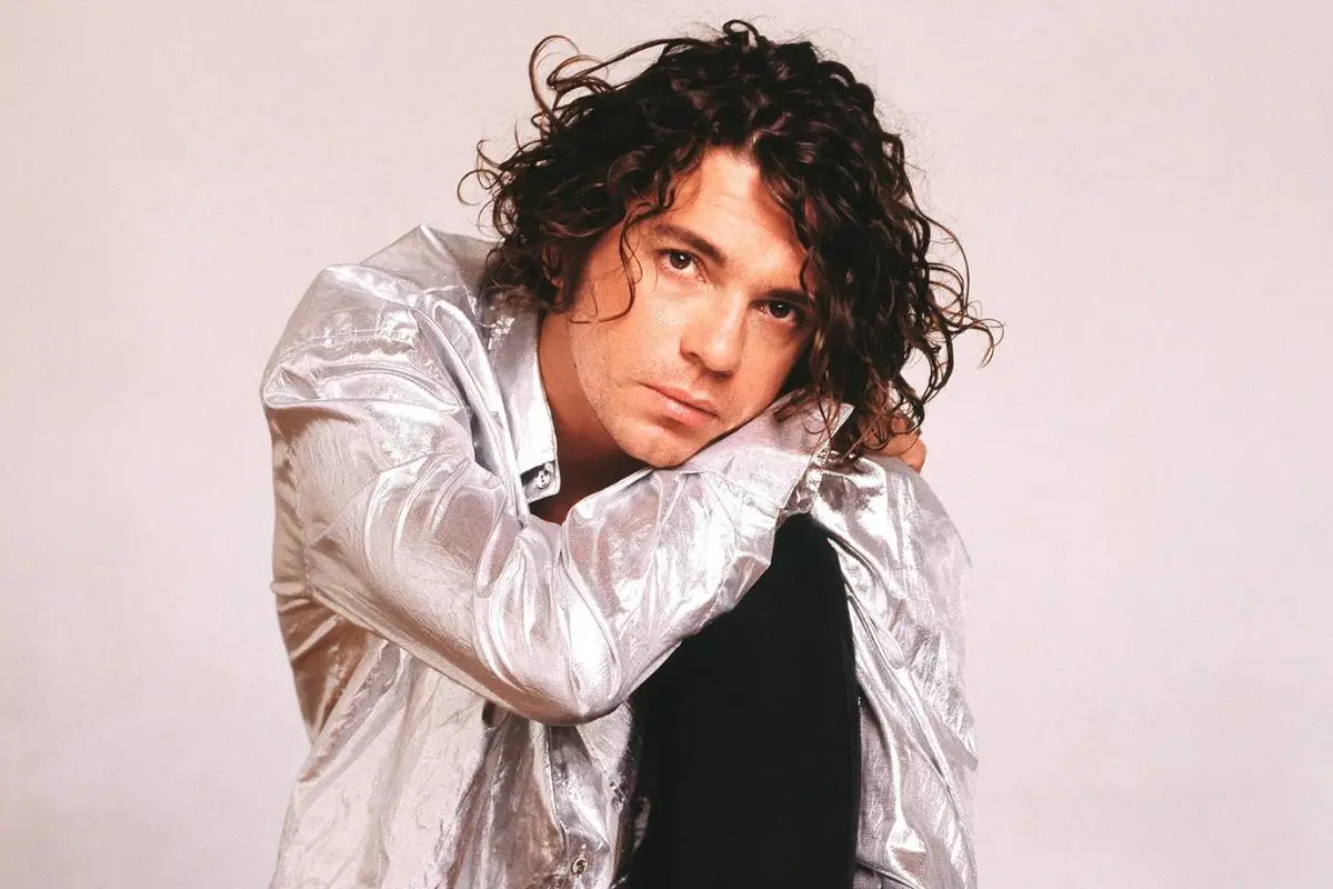 Michael Hutchence posing in a studio portrait, resting his head against his arm, with curly dark hair and an intense gaze, wearing a metallic silver jacket against a neutral background.