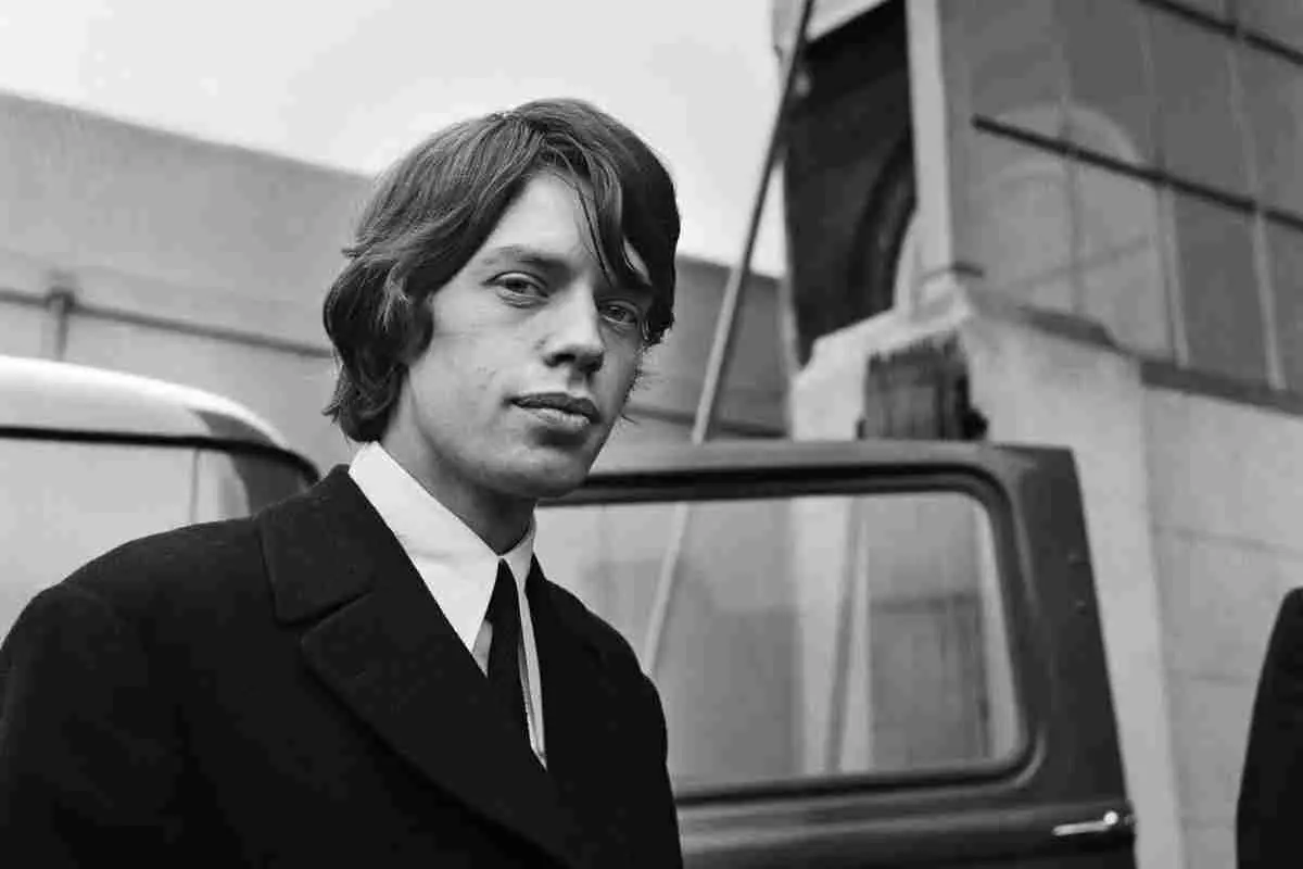 Mick Jagger standing outdoors in a black-and-white portrait, wearing a suit and tie.