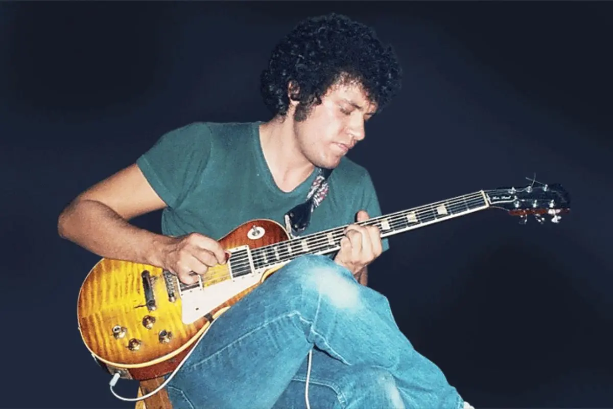 Mike Bloomfield seated while playing an electric guitar in a color performance photo, wearing a green T-shirt and jeans, captured mid-solo under stage lighting.