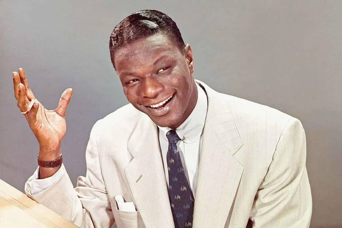 Smiling Nat King Cole in a pale suit jacket and patterned tie, seated and gesturing with his right hand while looking off-camera.