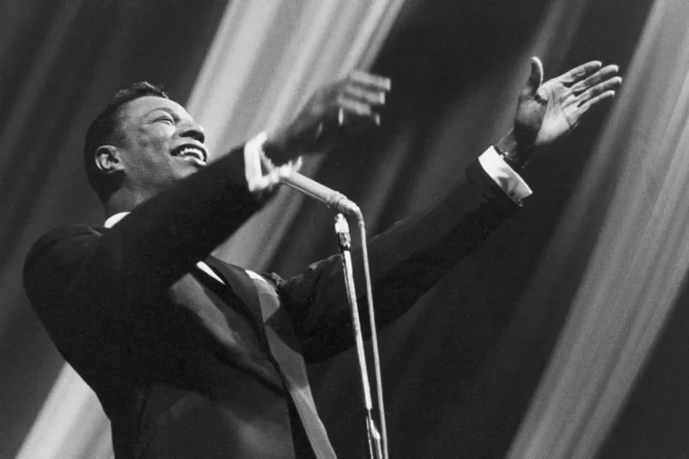 Nat King Cole on stage with arms outstretched, singing into a vintage microphone and wearing a dark suit.