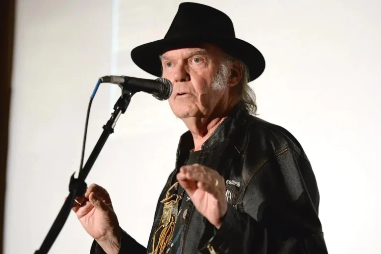 Neil Young speaking into a microphone, wearing a black hat and leather jacket.