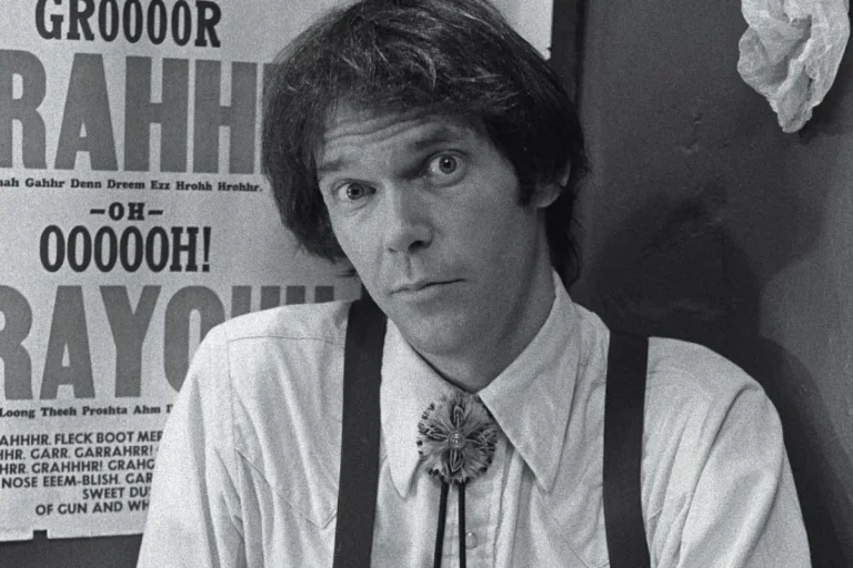 Neil Young vs the 5 Percent: Sound Quality, Fake Vinyl and “Dead” Rock & Roll Neil Young dressed in a collared shirt with suspenders stands indoors, looking directly at the camera with a humorous, wide-eyed expression in front of a poster filled with exaggerated text.