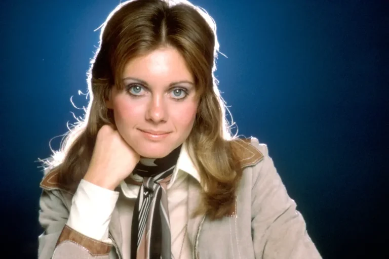 Olivia Newton-John at Eurovision 1974: The Awkward UK Gig in ABBA’s “Waterloo” Year Olivia Newton-John resting her chin on her hand against a blue backdrop, conveying poise, confidence, and a soft 1970s pop aesthetic.