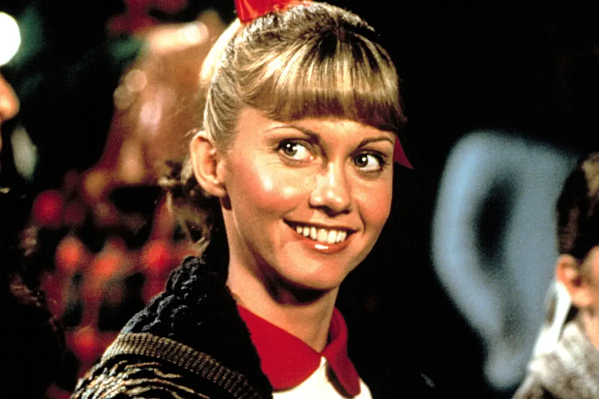 Olivia Newton John in a brightly lit scene, styled with a high ponytail and bangs, conveying optimism and the polished look of a classic musical film moment.