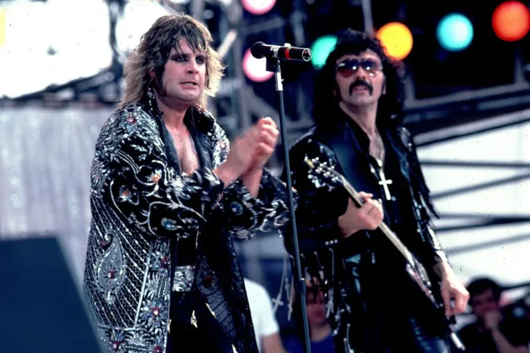 Ozzy Osbourne and Tony Iommi performing together on stage, with Ozzy clapping near a microphone stand and Tony playing guitar beside him, both wearing dark, embellished stage outfits under colorful concert lights.