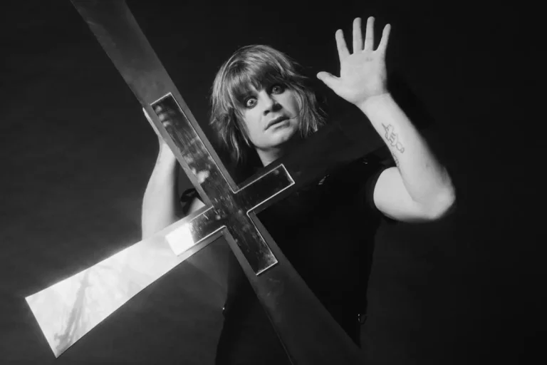 Ozzy Osbourne holding a large cross, raising one hand with a dramatic, wide-eyed expression.