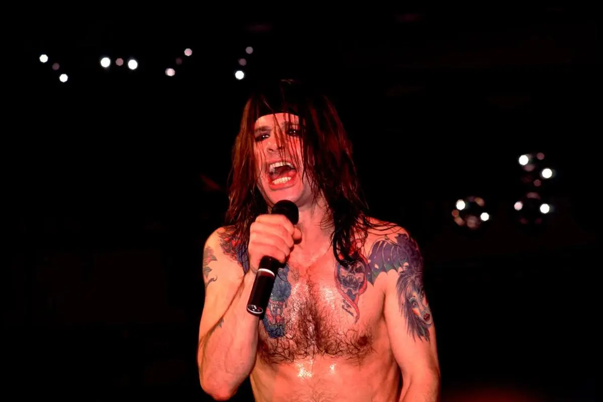 Ozzy Osbourne with long hair grips a microphone mid-performance, singing intensely under dark stage lighting.