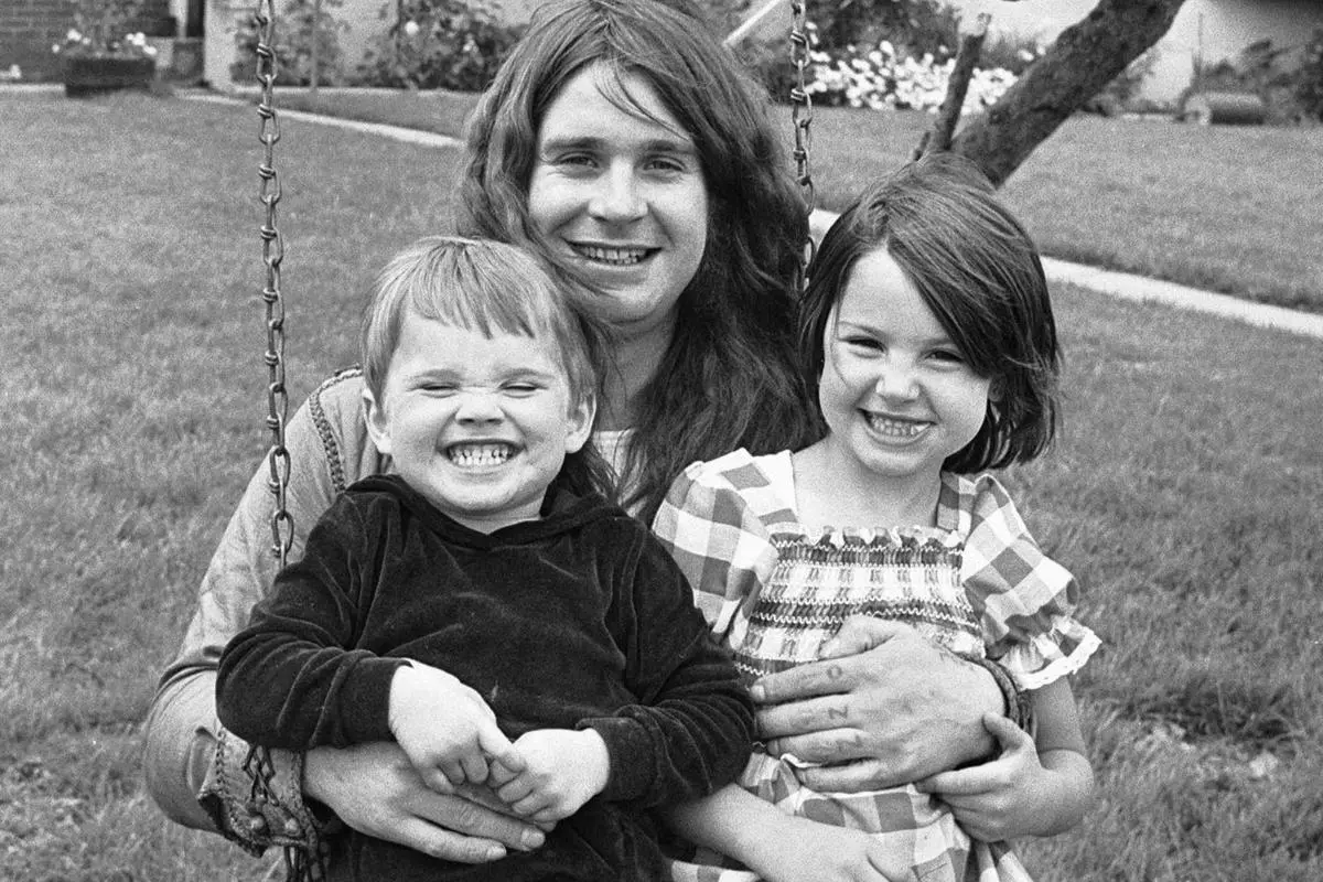 Ozzy sitting on a swing with two young children on his lap, all three smiling broadly at the camera.