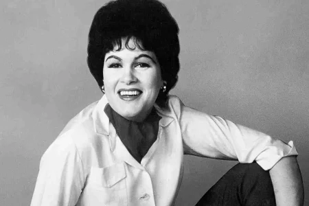 Patsy Cline posing in a black-and-white portrait, smiling warmly while seated, wearing a light-colored blouse with her hair styled in a classic bouffant.