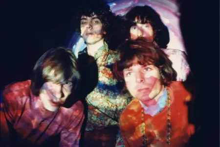 Pink Floyd band members in a psychedelic 1960s portrait with colorful projected patterns on their faces.