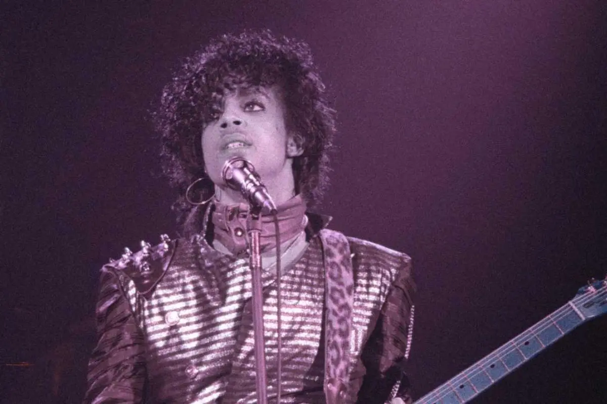 Prince singing and playing guitar under purple stage lighting.