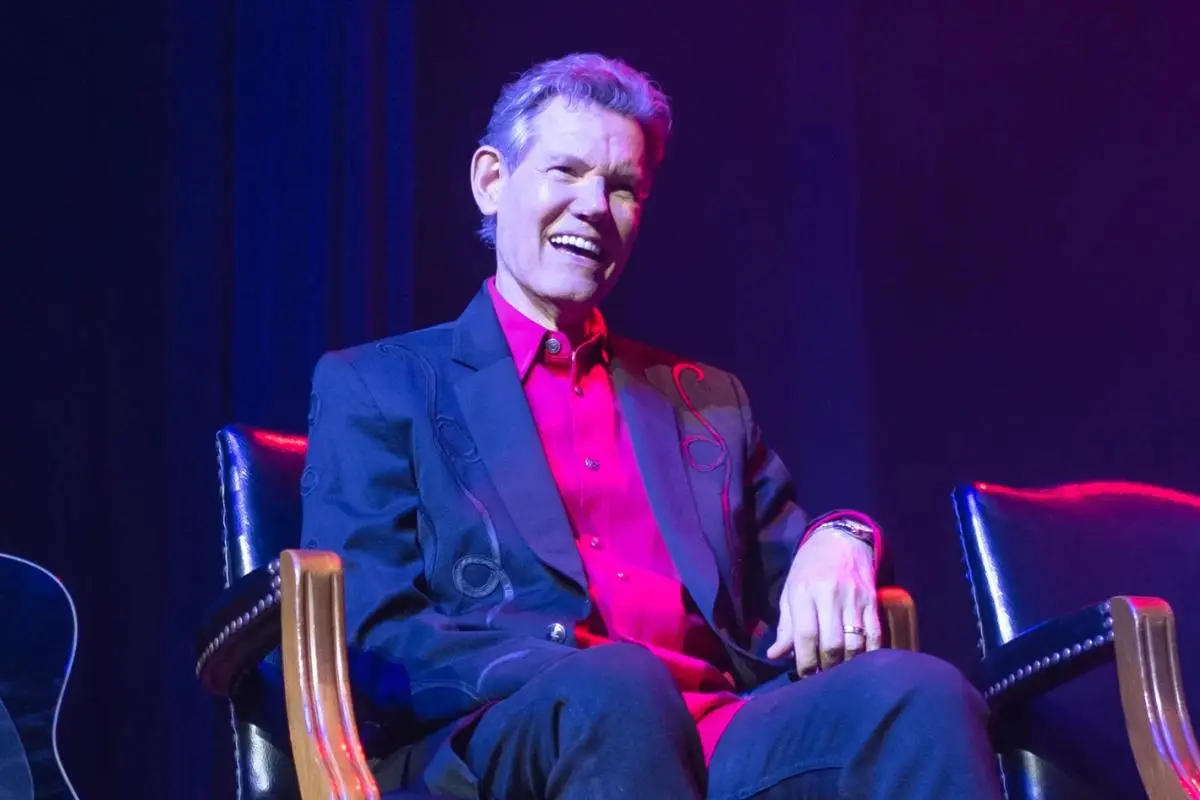 Randy Travis smiling while seated on stage under purple and blue lighting.