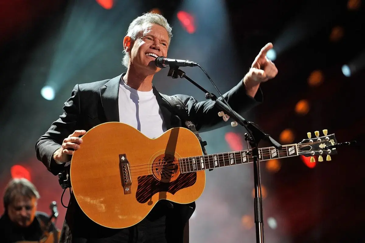Randy Travis performing live on stage, singing and pointing while playing an acoustic guitar.