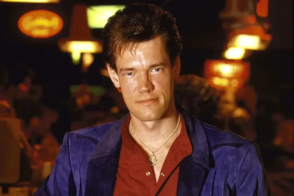 Portrait of Randy Travis wearing a red shirt and blue jacket, seated indoors with warm lighting.