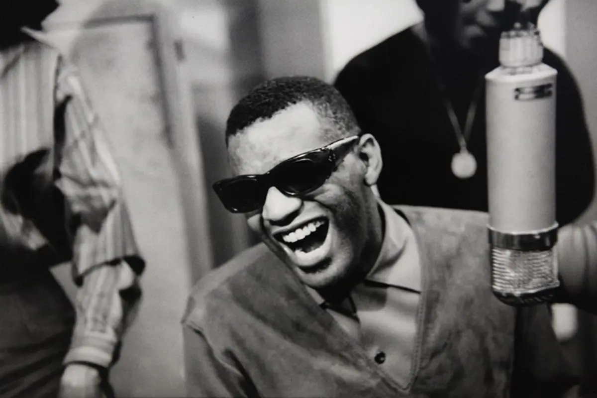 Black-and-white photo of Ray Charles laughing while singing into a studio microphone, wearing sunglasses.