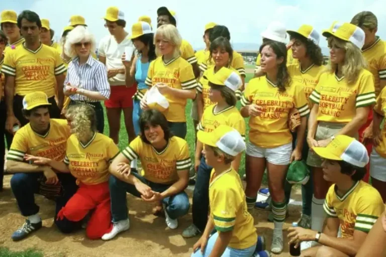 Riviera Softball team in matching yellow softball jerseys gather outdoors on a field, smiling and talking during a casual event.