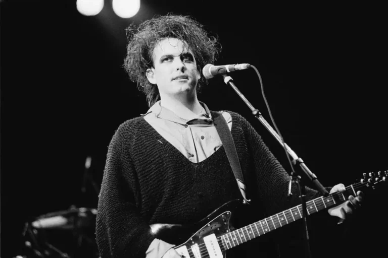 Robert Smith singing into a microphone onstage, wearing a loose sweater with tousled hair.