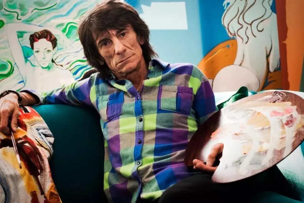 Ronnie Wood seated on a couch holding painter’s palettes in a colorful art studio.