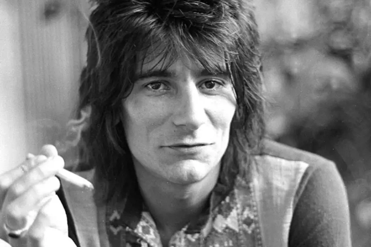 Black-and-white portrait of Ronnie Wood with shaggy hair, holding a cigarette and looking toward the camera.