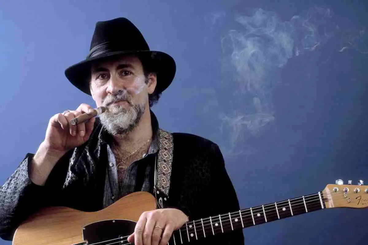 Roy Buchanan holding a guitar and smoking a cigar, wearing a black hat against a blue background