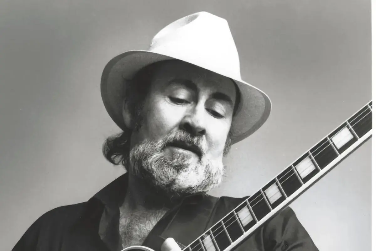 Roy Buchanan wearing a white hat looks down at his instrument with a gentle expression, his beard softly framing his face.