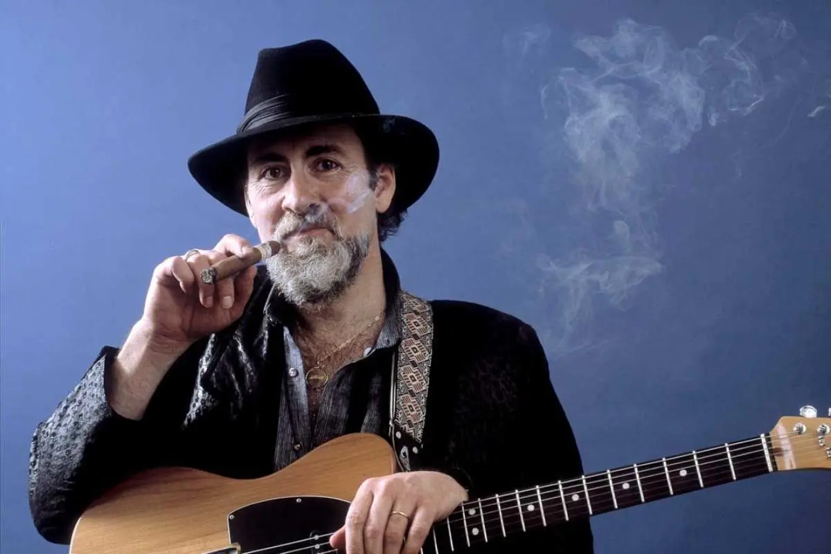 Roy Buchanan in a dark hat holds a lit cigar while resting his hand on an electric guitar, smoke curling around him.