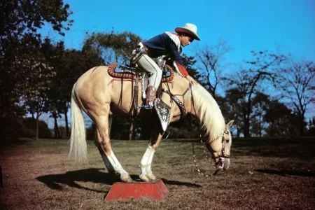 Roy Rogers riding his horse outdoors in a color photo, dressed in full western attire, leaning forward in the saddle during a trick-riding or training moment.