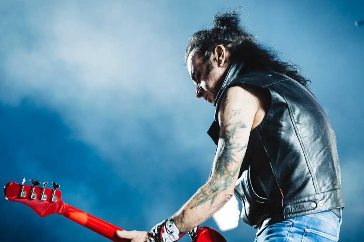 Simon Gallup performing on stage playing a red bass guitar, wearing a sleeveless leather vest.