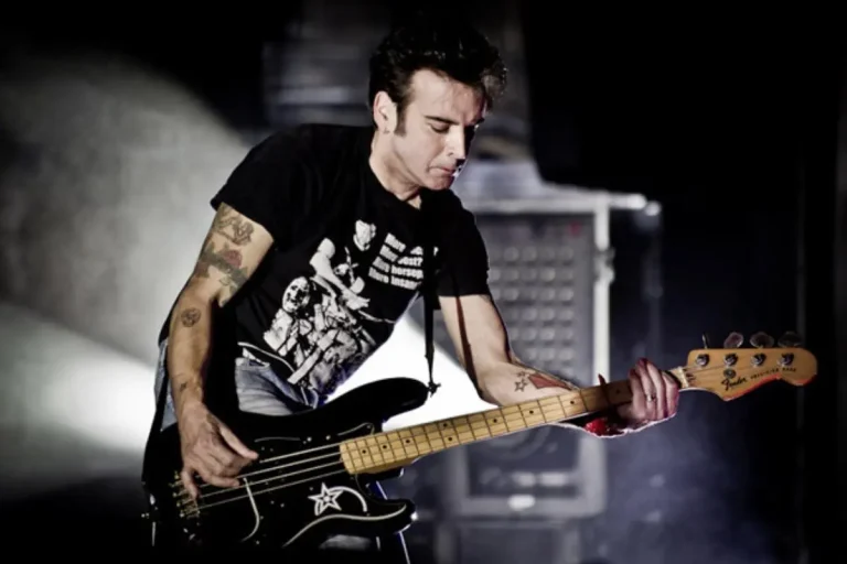 Simon Gallup playing bass guitar live under stage lighting, wearing a black T-shirt.