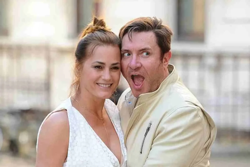 Simon Le Bon and Yasmin Le Bon posing together outdoors, smiling closely at the camera, with Simon wearing a light-colored jacket and Yasmin wearing a white sleeveless dress.