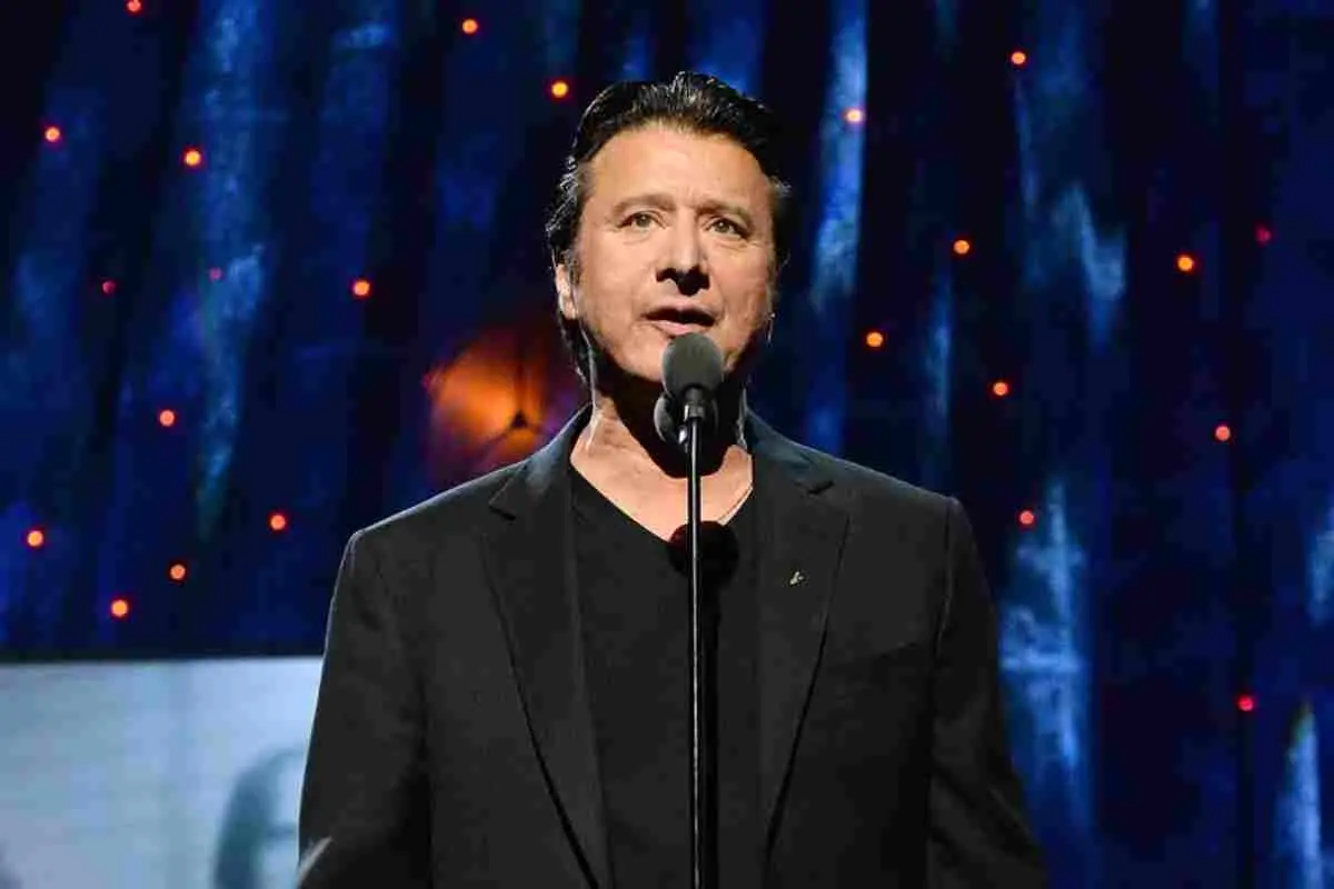 Steve Perry speaking into a microphone on stage, wearing a dark suit.