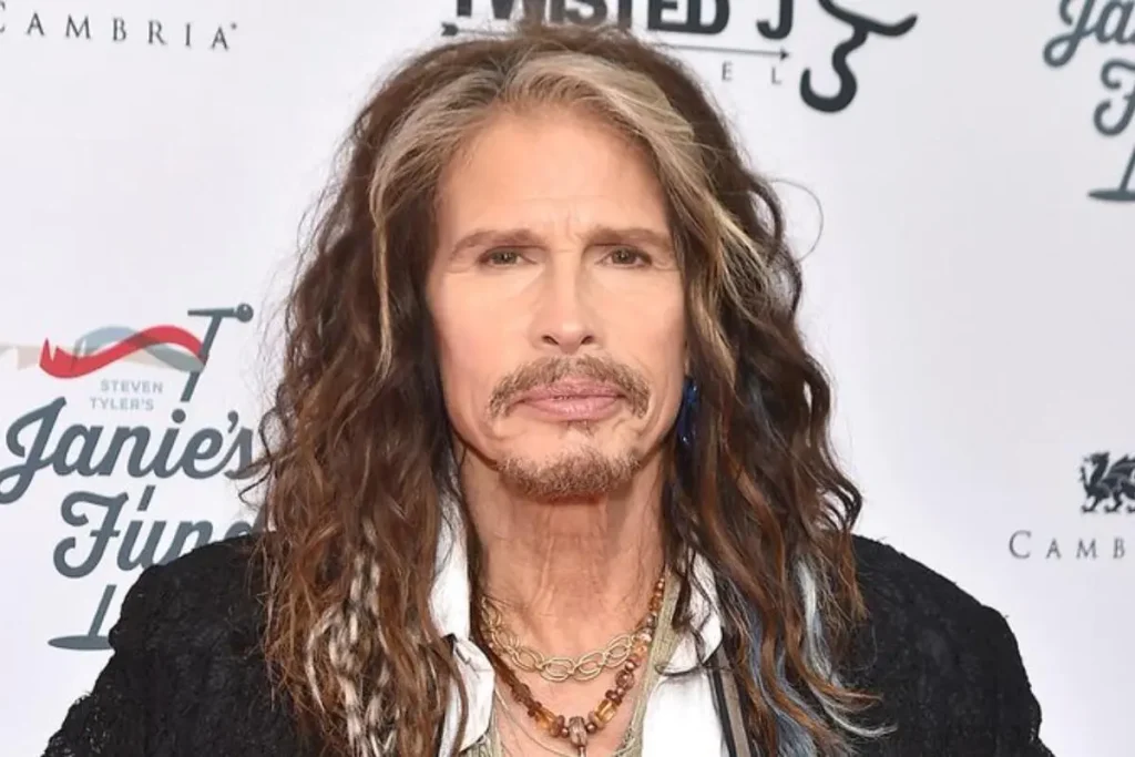 Steven Tyler posing at an event, wearing layered necklaces and a black jacket.