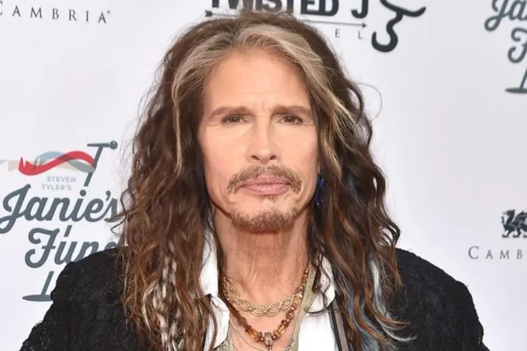 Steven Tyler posing at an event, wearing layered necklaces and a black jacket.
