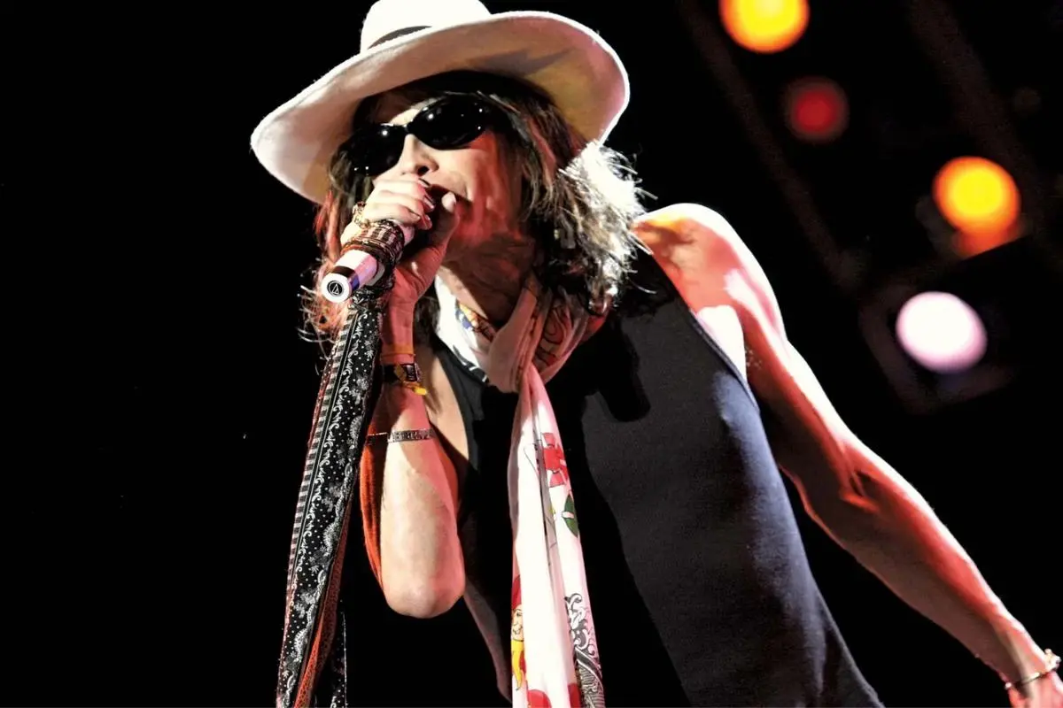 Steven Tyler on stage wearing a white hat and sunglasses, singing into a microphone.
