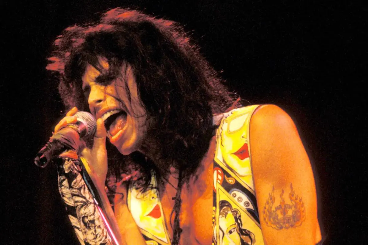 Steven Tyler mid-performance, mouth open while singing under warm stage lights.