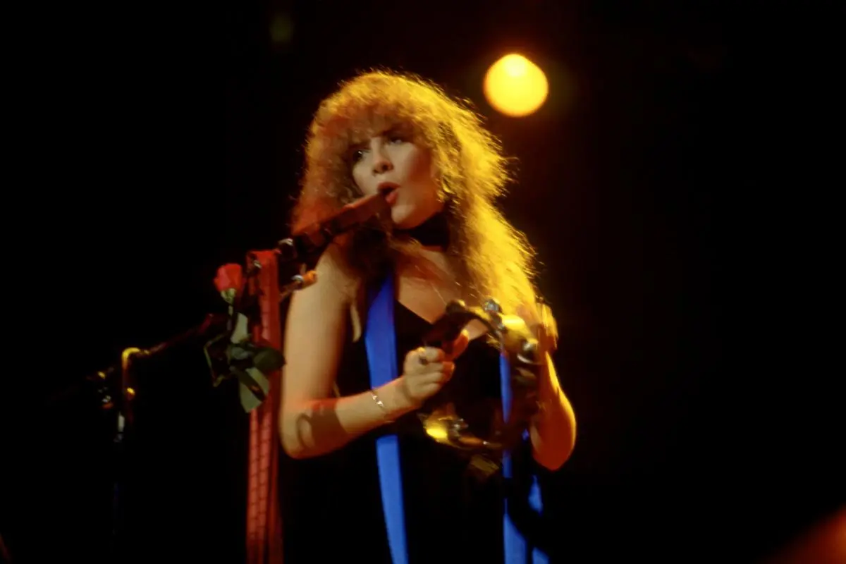 Stevie Nicks singing into a microphone while playing tambourine under warm stage lights.