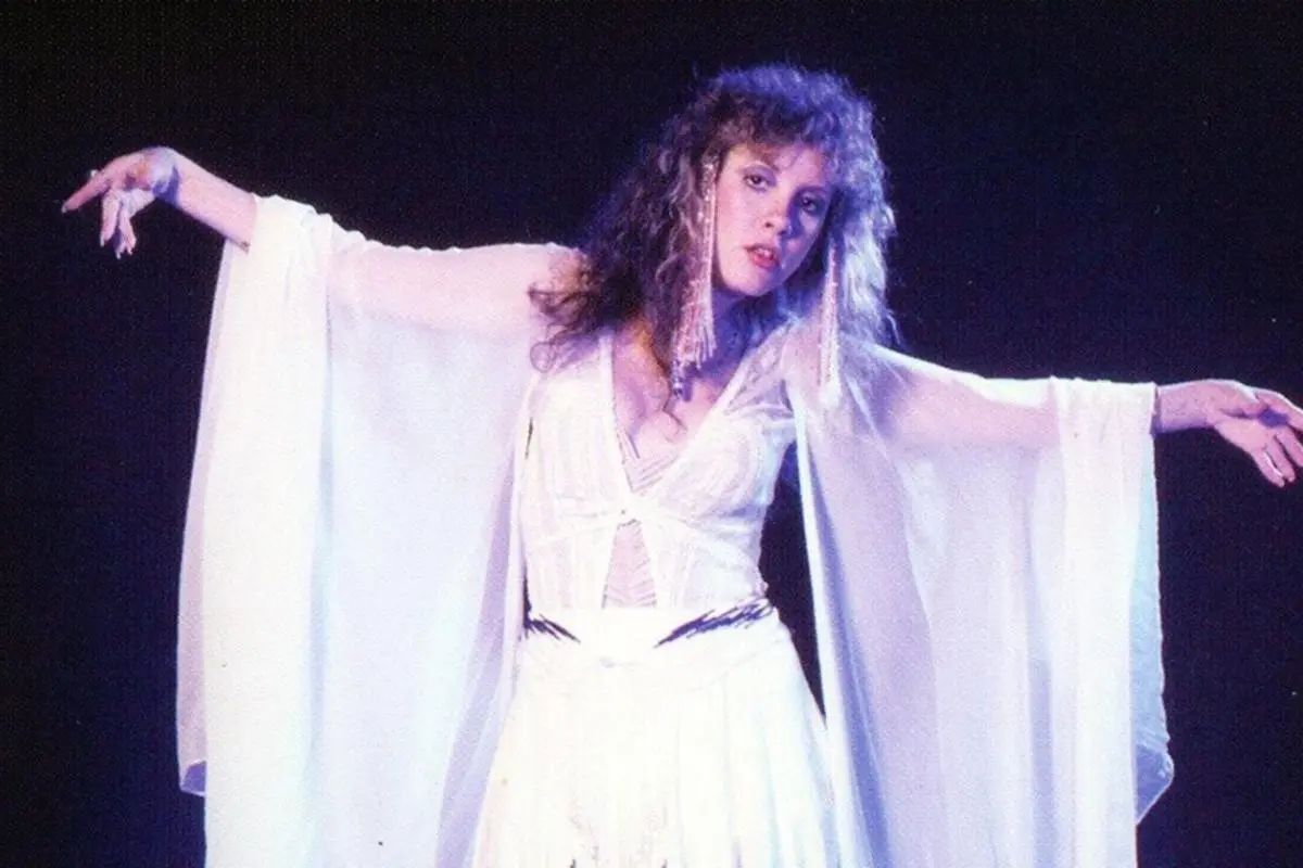 Stevie Nicks performing onstage with arms outstretched in a flowing white outfit.