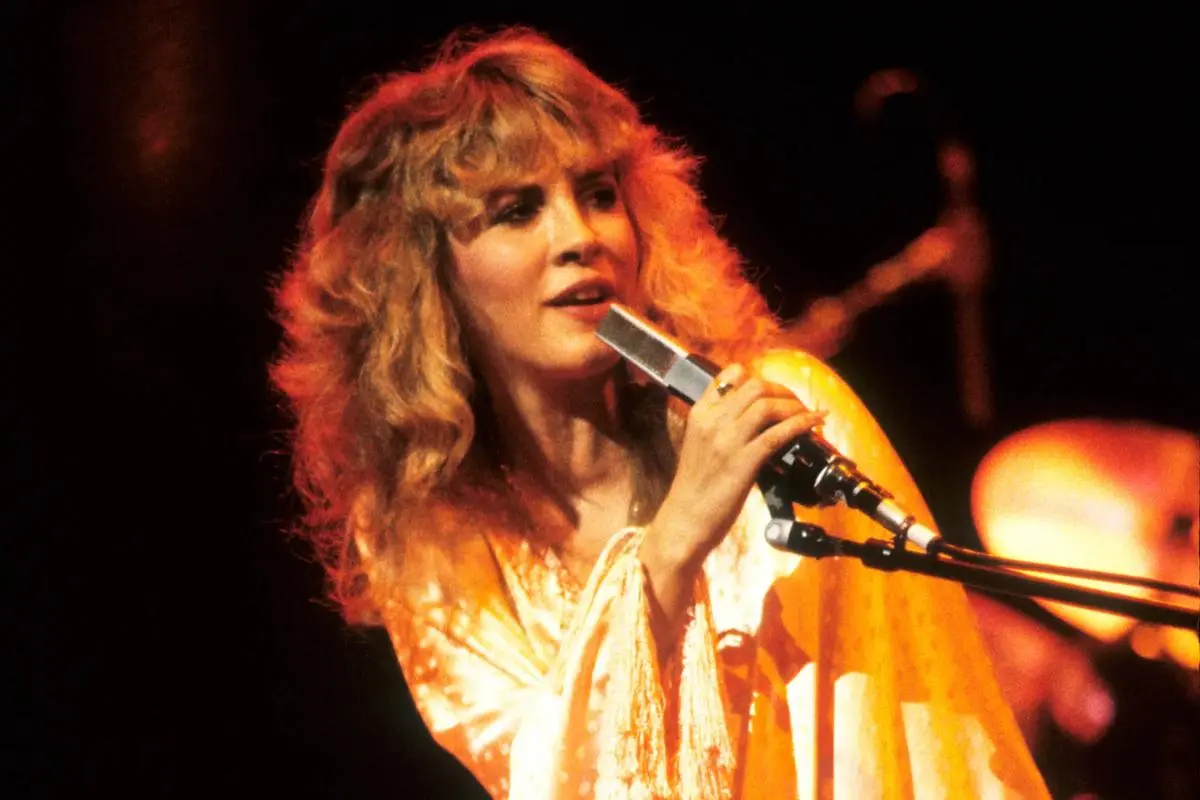 Stevie Nicks singing into a microphone on stage, wearing a flowing golden outfit, with curly blonde hair illuminated by warm orange stage lights.