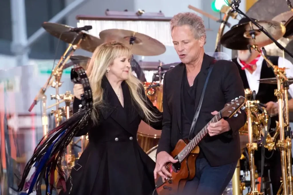 Stevie Nicks and Lindsey Buckingham performing together on stage, with Nicks holding a tambourine and Buckingham playing guitar, surrounded by drums and concert lighting.
