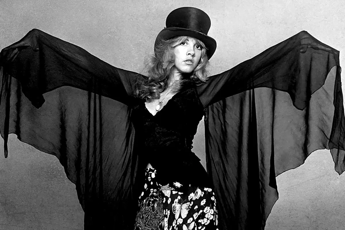 Stevie Nicks in a top hat spreads her sheer, flowing sleeves like wings while posing confidently in a dramatic, vintage-style outfit.
