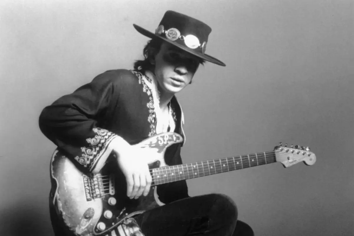 Stevie Ray wearing a wide-brim hat, seated and holding an electric guitar.
