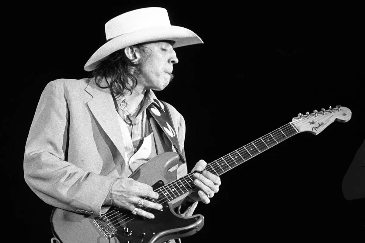 Stevie Ray Vaughan playing electric guitar on stage, wearing a white cowboy hat in a black-and-white performance photo.