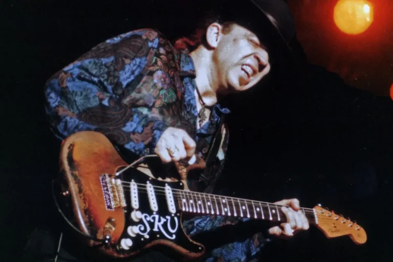 Stevie Ray Vaughan in a patterned shirt and wide-brimmed hat plays an electric guitar intensely onstage, his face tightened with emotion under dramatic lighting.