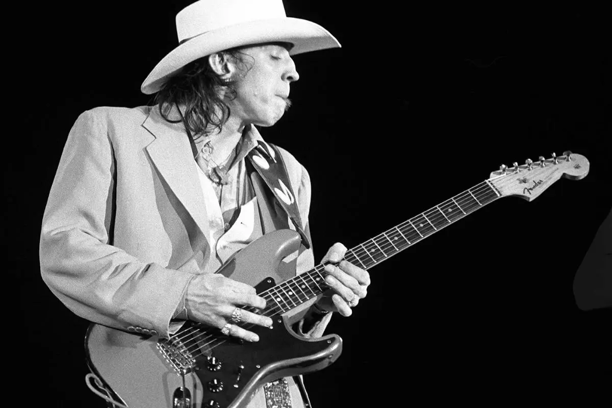 Stevie Ray in a suit and cowboy hat playing an electric guitar onstage.
