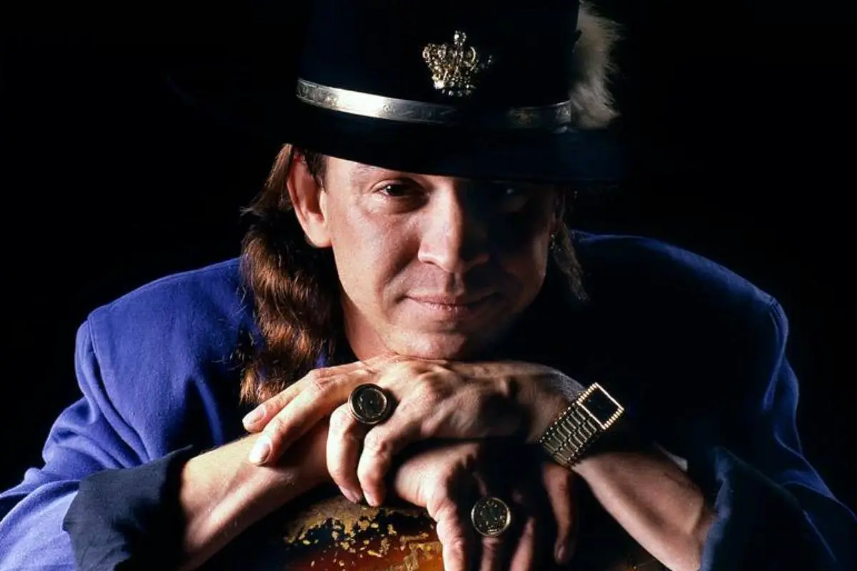 Portrait of Stevie Ray Vaughan wearing a hat and rings, resting his hands on a guitar.
