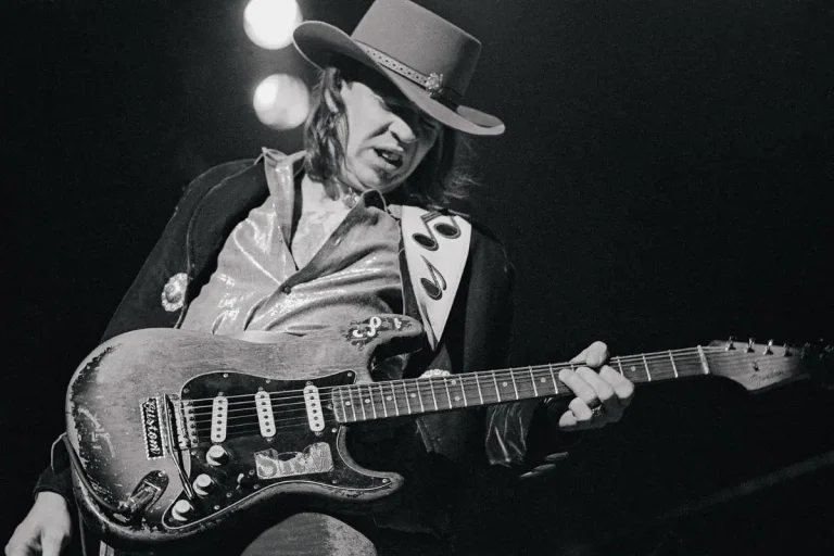 Stevie Ray Vaughan performing live on stage, leaning into his guitar solo while wearing a wide-brimmed hat.