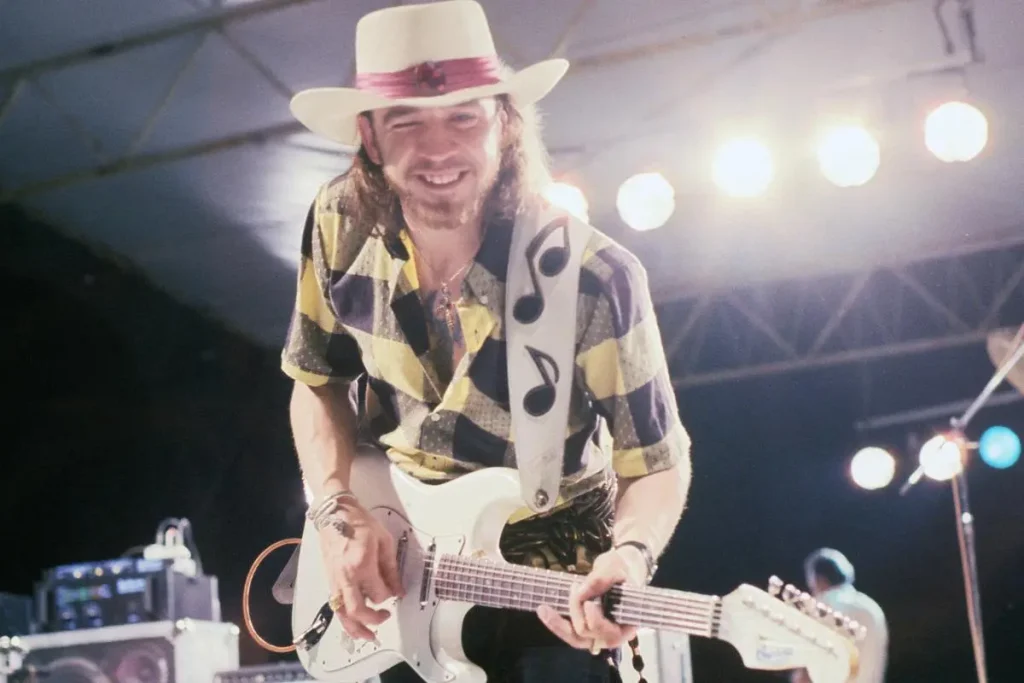 Stevie Ray in a hat playing an electric guitar under bright stage lights.