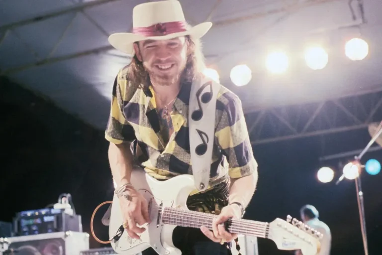 Stevie Ray in a hat playing an electric guitar under bright stage lights.